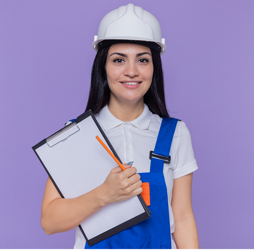 young builder woman construction uniform safety helmet holding clipboard pencil looking front happy positive smiling confident standing purple wall 1